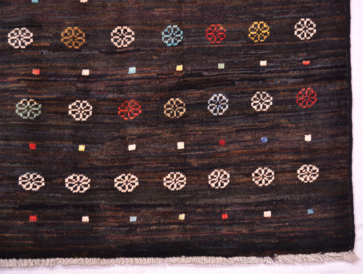 A close-up of a dark brown handwoven Afghan Gabbeh rug with a textured surface, featuring rows of geometric medallion motifs in cream, red, blue, yellow, and green, finished with a fringed edge.