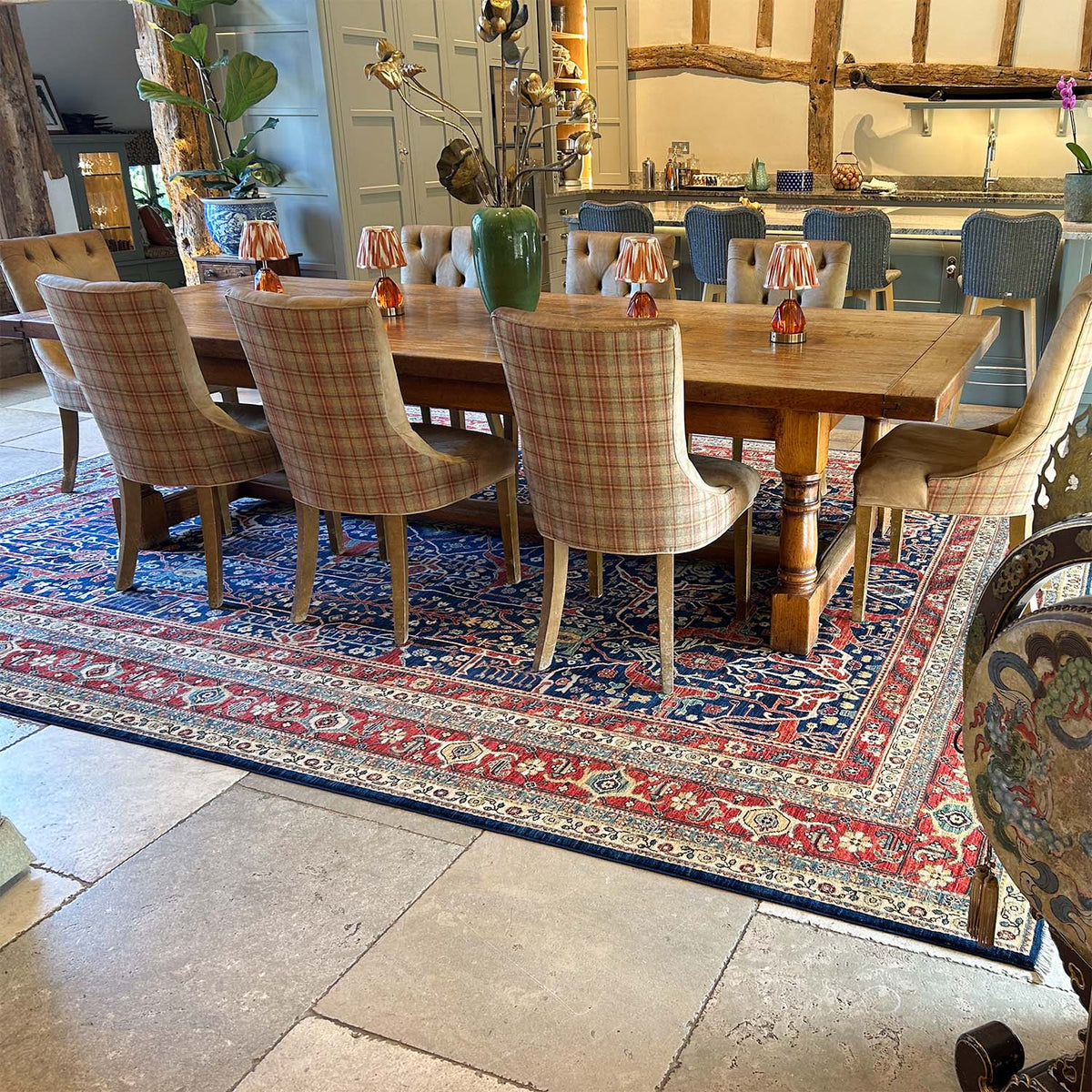 A rustic wooden dining table surrounded by six plaid upholstered dining chairs, set on a large traditional patterned hand-knotted fine Afghan Garous Carpet with rich colours in a dining room.