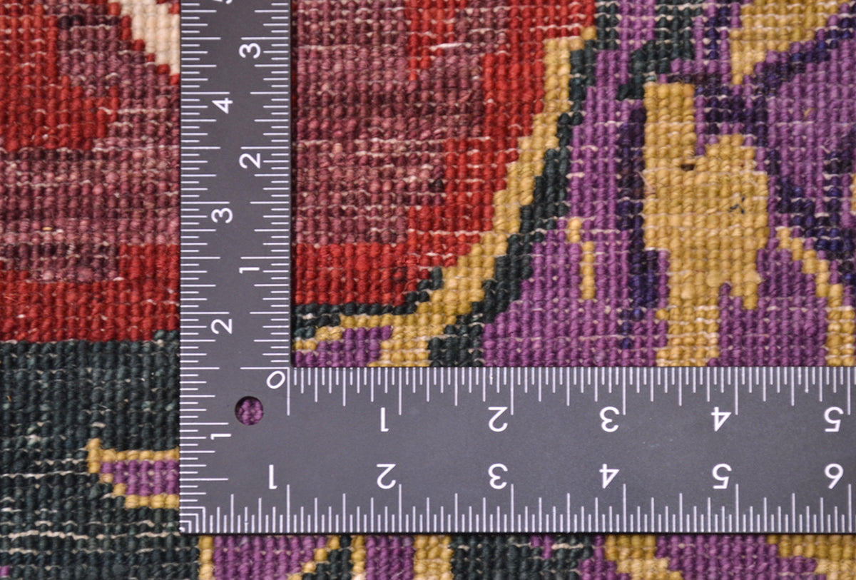 Close-up of the hand-knotted back of a patterned Afghan Gul Ferang rug with a dark gray metal ruler placed vertically and horizontally to show scale, featuring red, purple, yellow, and dark green woven details.