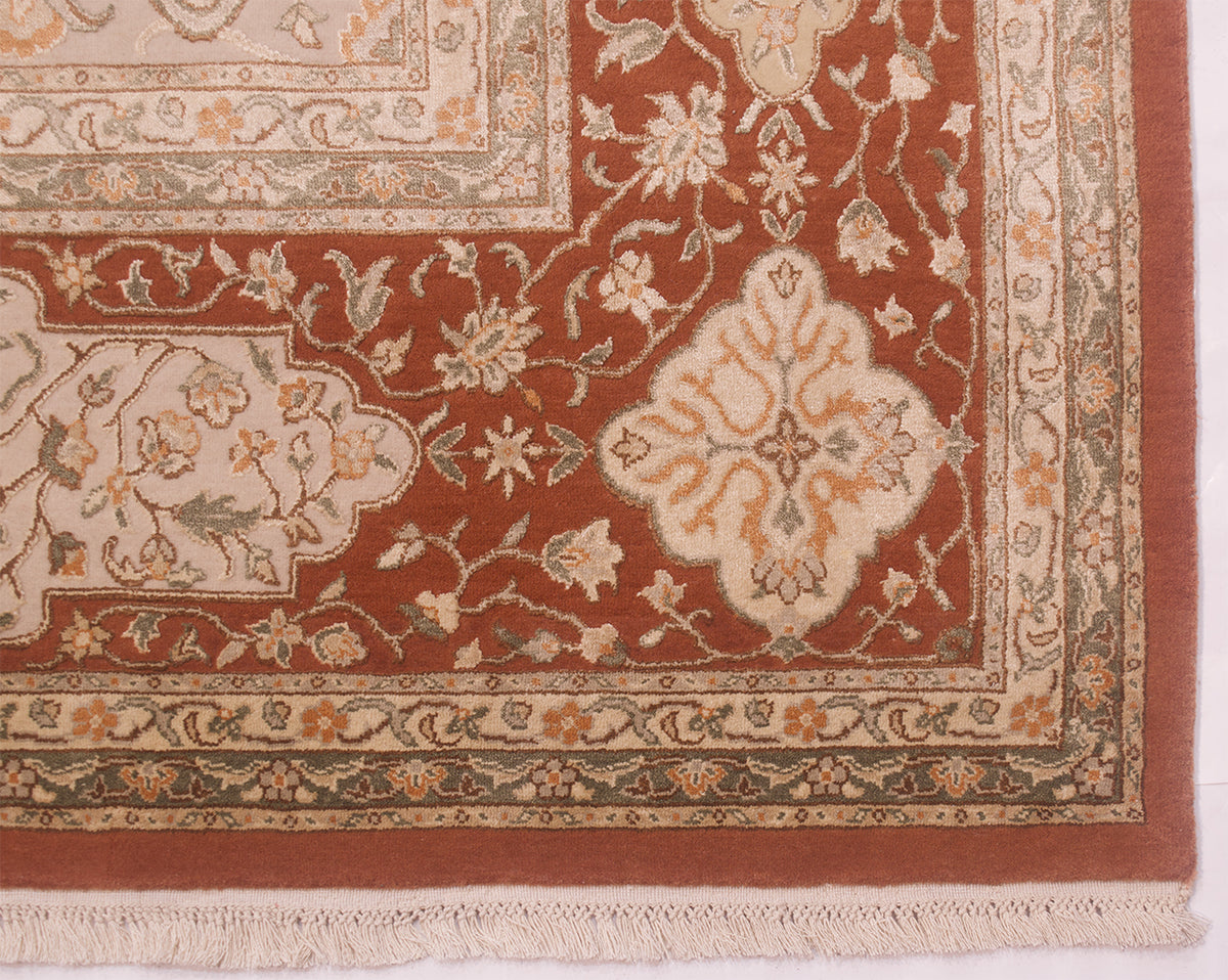 A close-up of an ornate area rug corner showing a rust red field, cream central medallion, beige floral motifs, and a decorative border with fringe along the bottom edge.