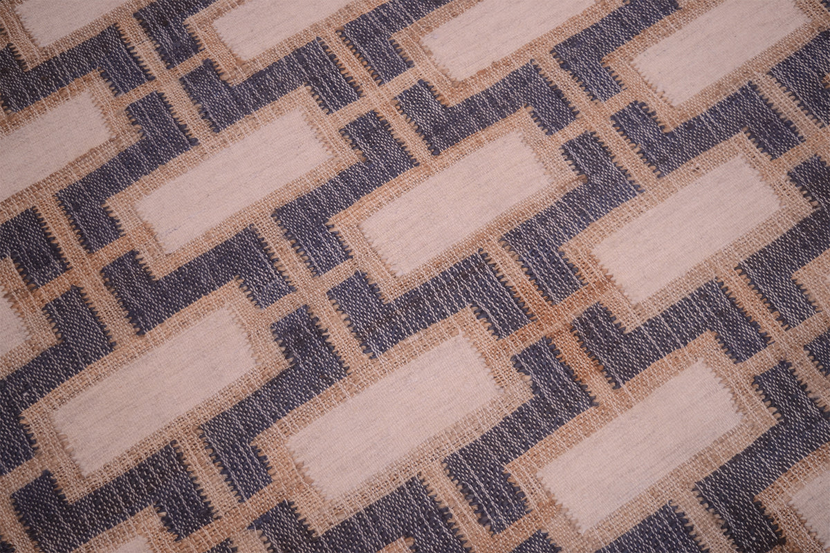 A close-up of a woven rug or textile with a geometric pattern of beige rectangles outlined in tan, separated by dark navy zigzag lines.