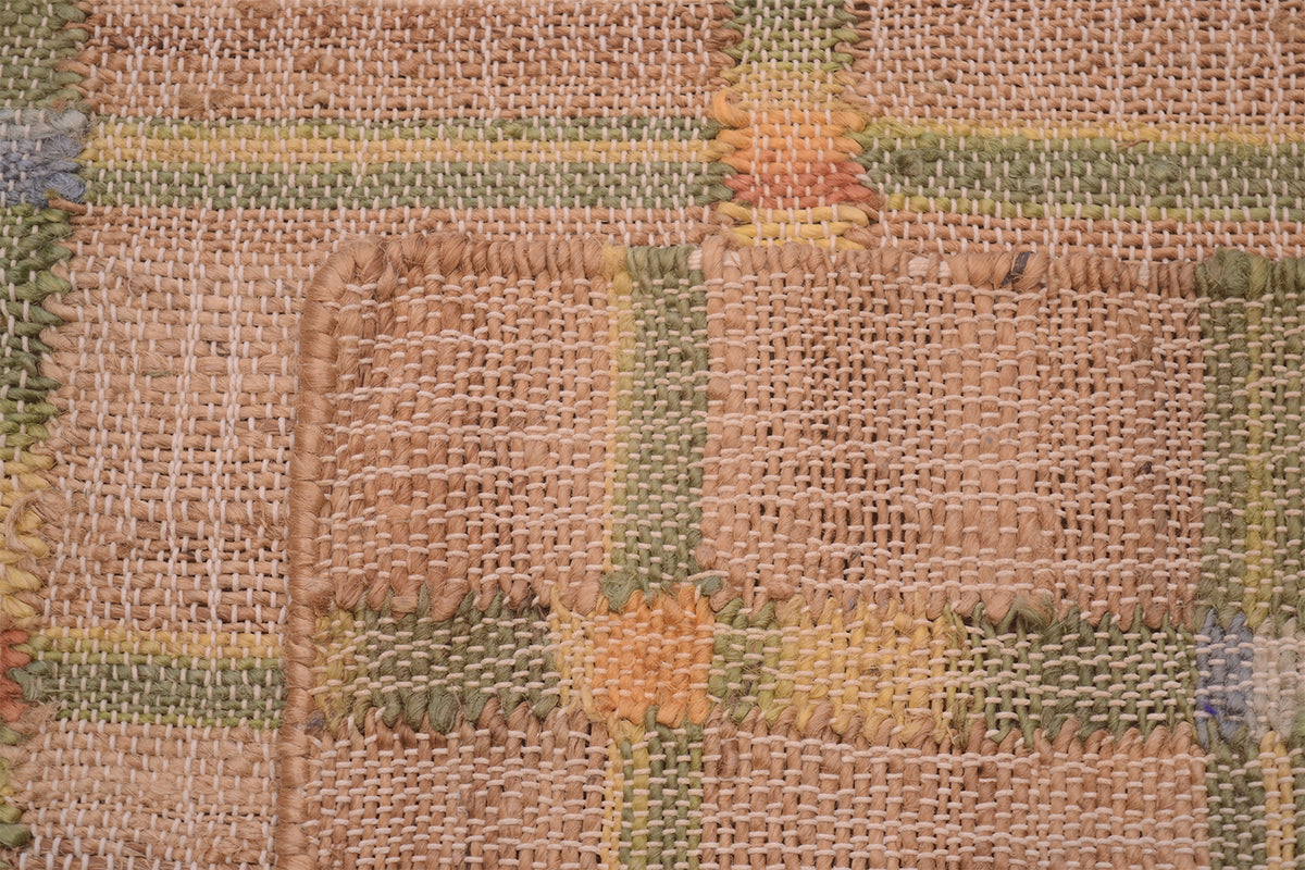 A close-up of a rustic woven textile or rug with a patchwork design, featuring natural tan burlap-like sections and stitched blocks in muted green, yellow, and orange.