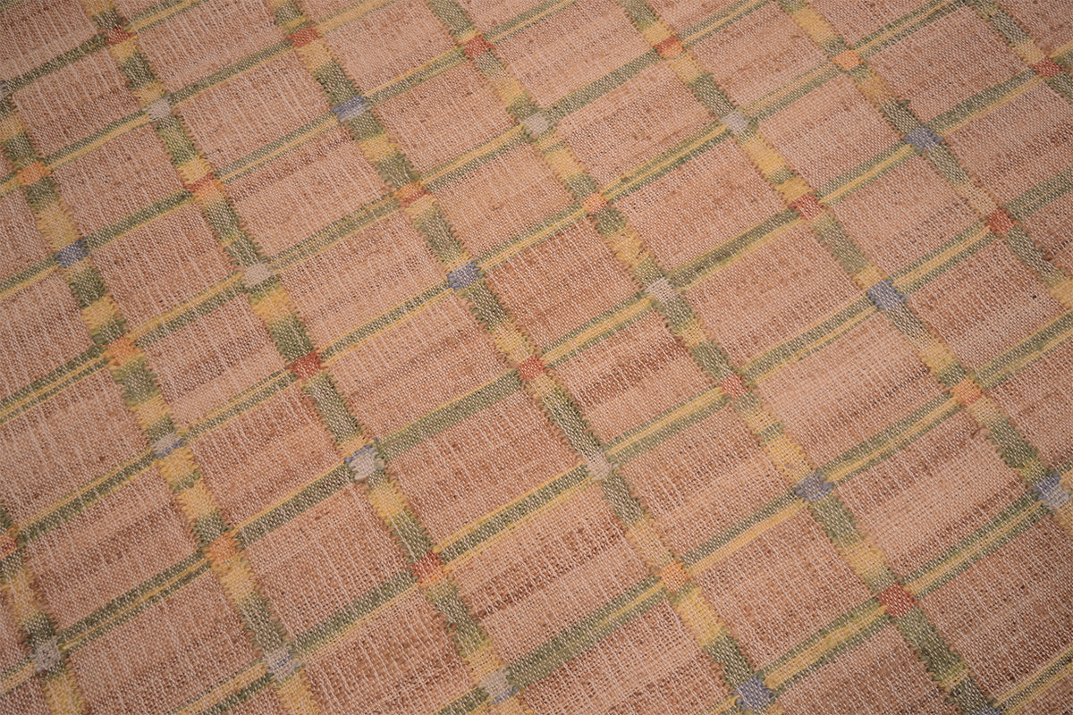 A close-up of a woven textile or rug with a tan and beige base, overlaid with a repeating plaid pattern in muted yellow, olive green, and small blue accents.