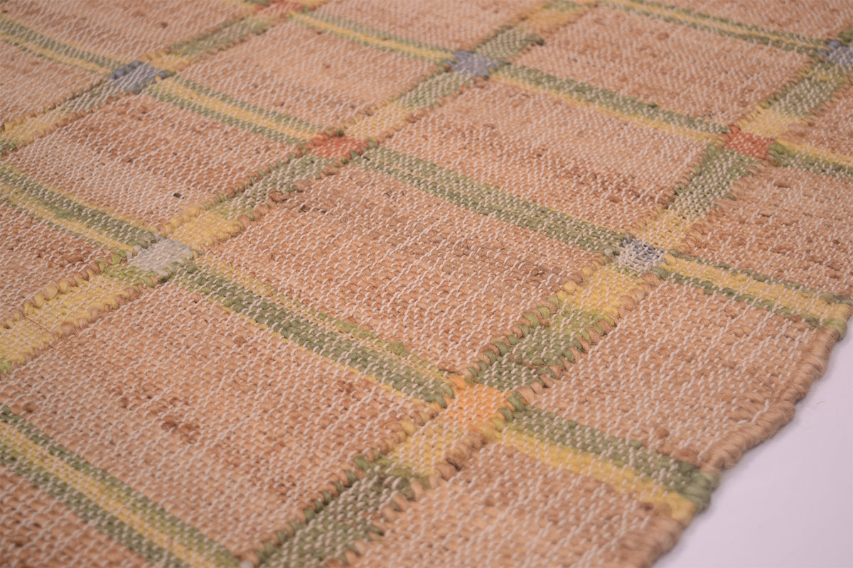 A close-up of a woven textile, likely a rug or mat, featuring a warm beige base with a plaid pattern in muted green, yellow, and orange.