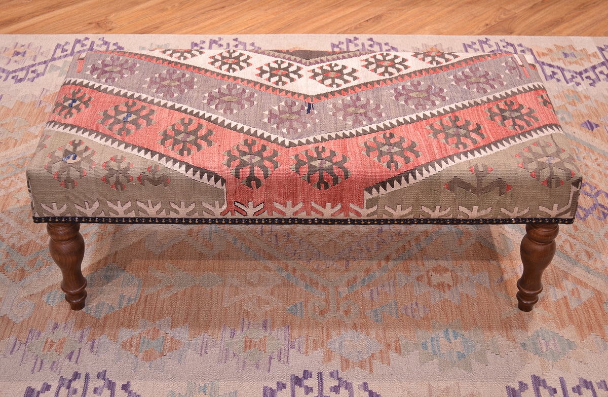 Vintage Kilim Bench with geometric kilim upholstery in soft terracottas. creams, light greens. soft greys and beige natural cream and chestnut brown stylized hooked mini medallions and motifs. Dark borwn wooden legs. and beaded edging. Kilimbench resting on a soft wash Afghan kilim with muted colours.