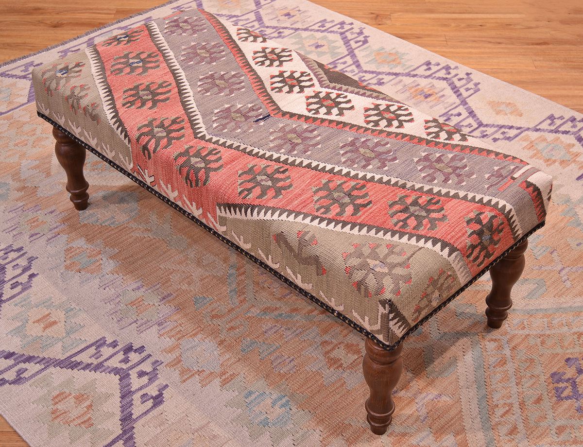 View of vintage Anatolian kilim in soft greens, beige and terracottas with beige/cream detailing. Resting on a sfot ws afghan kilim and wooden floor. Dark wooden legs. Vintage turkish kilim used for upholster kilim bench.
