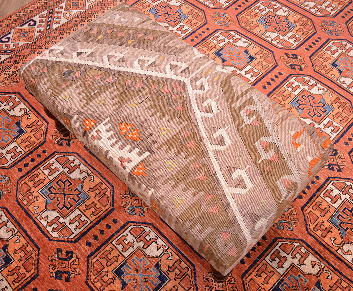 Top overall view of long bench stool covered with soft muted tones of vintage turkish kilim with cream, light olive green, coral pinks and hints of orange. Kilim bench stool placed on a terracotta Afghan Ersari carpet with fil pai design.