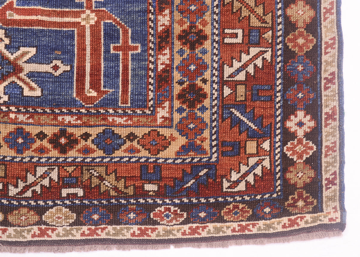 A close-up of a traditional patterned Caucasian Karagashli rug corner showing a blue field, red and brown geometric motifs, and beige and navy borders.