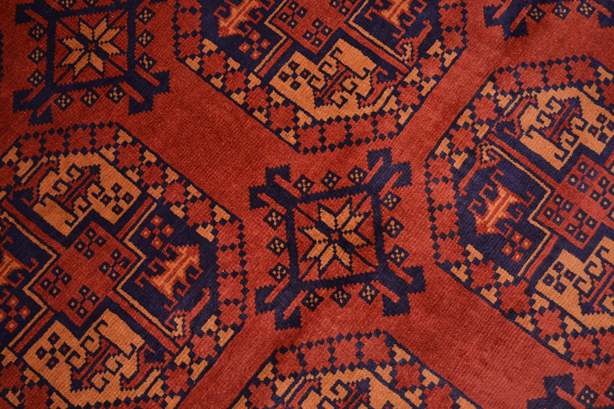 A close-up of a patterned Afghan Kargahi rug with a rust red base, dark navy geometric motifs, and soft orange accents.