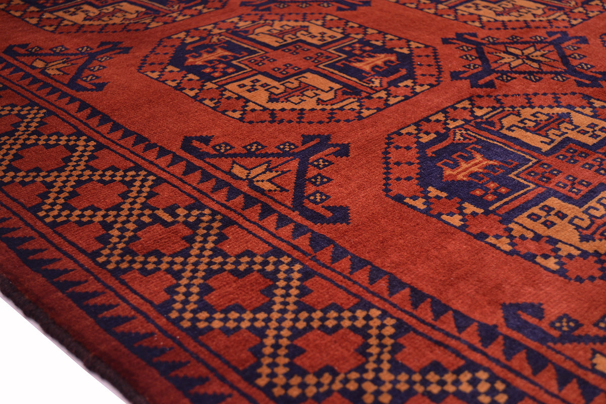 A close-up of a traditional patterned Afghan rug with a rust-red field, dark navy blue geometric motifs, and soft orange accents.