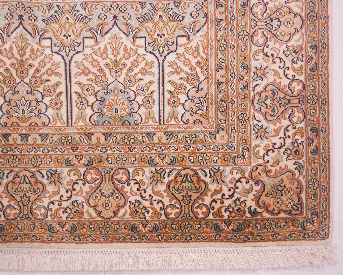 A close-up of an ornate Kashmir silk area rug corner, showing intricate floral patterns in beige, rust orange, and navy blue with white fringe.