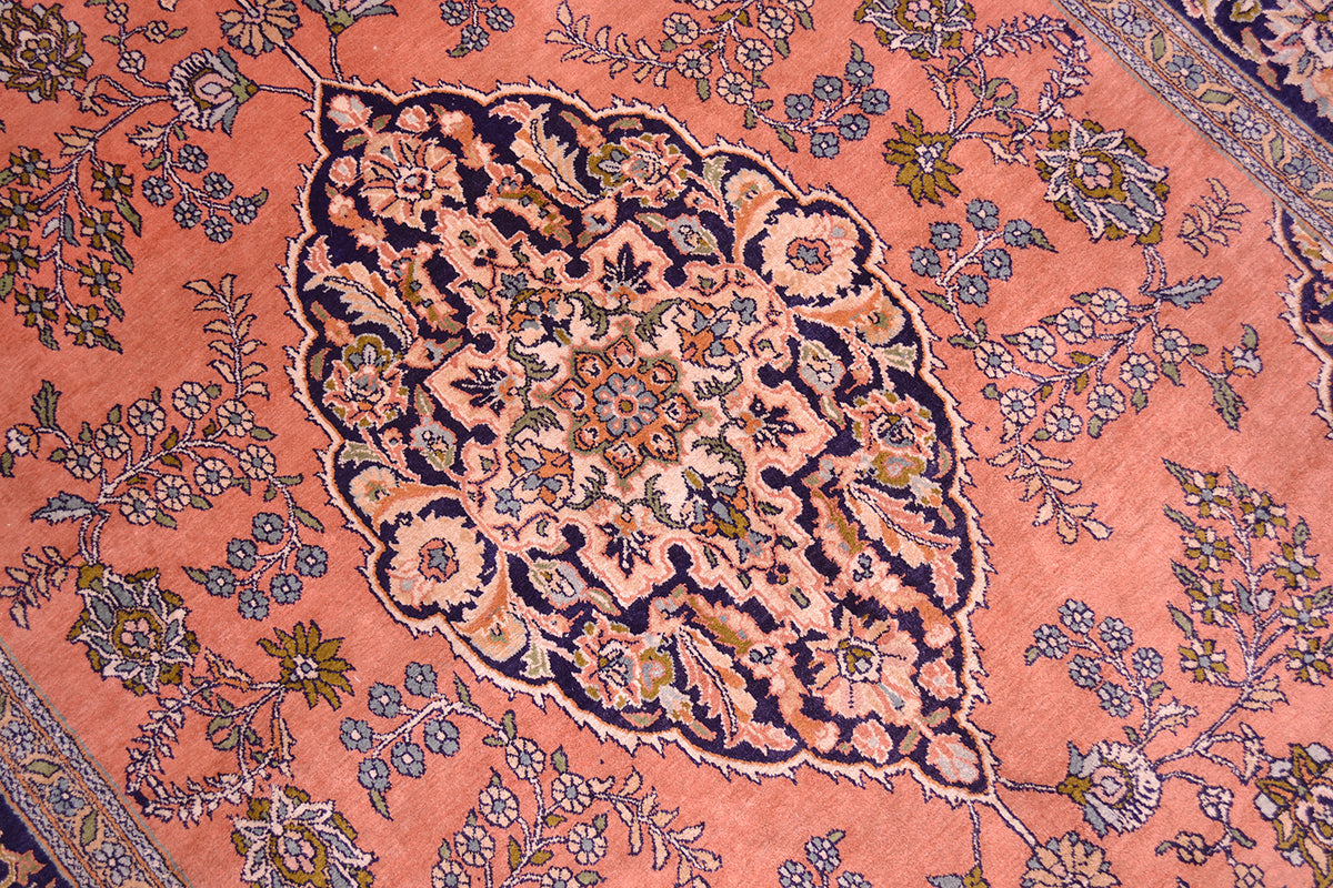 A close-up of a traditional old silk Kashmir rug featuring a large central teardrop-shaped medallion in navy blue, beige, and gold, surrounded by floral motifs on a warm pink field.