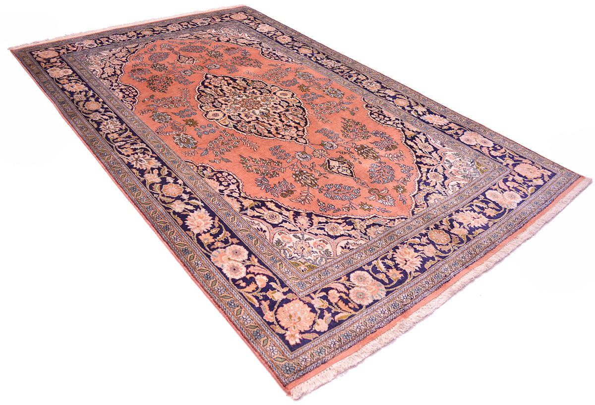 A traditional Silk Kashmir rug with a dusty pink center, navy blue borders, and beige floral motifs, shown from an elevated angle on a white background.
