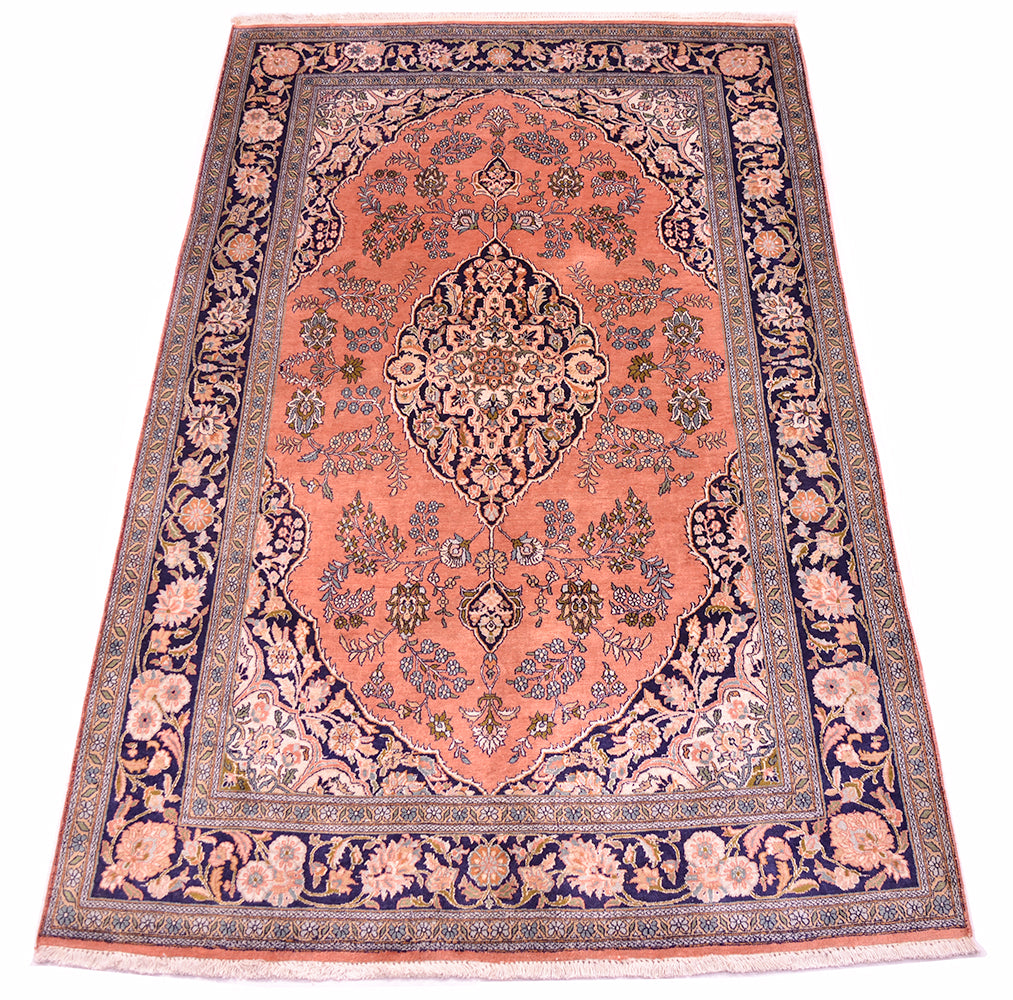 A silk Kashmir rug with a coral pink field, navy blue borders, and intricate floral motifs in beige, green, and blue, shown flat against a white background.