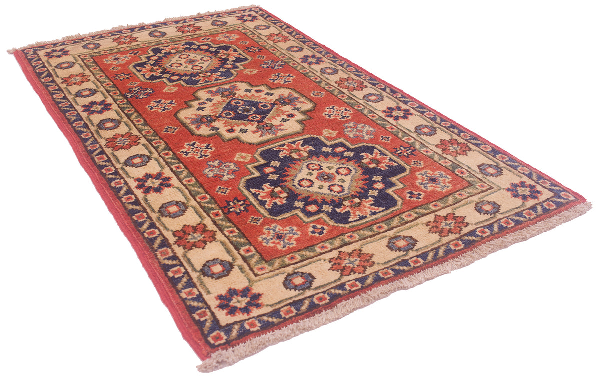 A rectangular small size handmade Kazak Mat with a red field, beige borders, and navy blue medallions, shown at an angle on a white background.