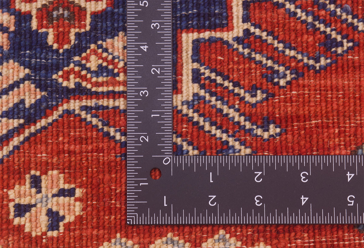 A close-up of the hand-knotted weave of a rust-red Afghan fine Kazak Rug with a dark metal ruler placed over it to show scale of the knotting. The rug has a red-orange base with navy blue and beige geometric and floral motifs.