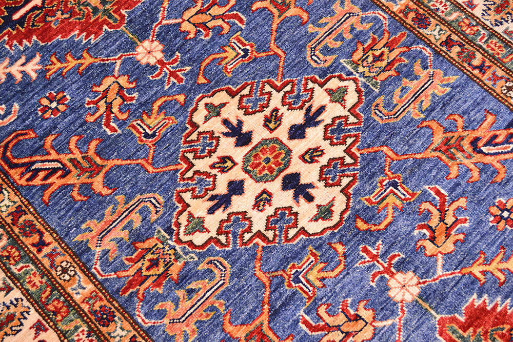 A close-up of a handmade Fine Kazak wool Rug with a blue field, cream central medallion, and orange, red, and green floral motifs.