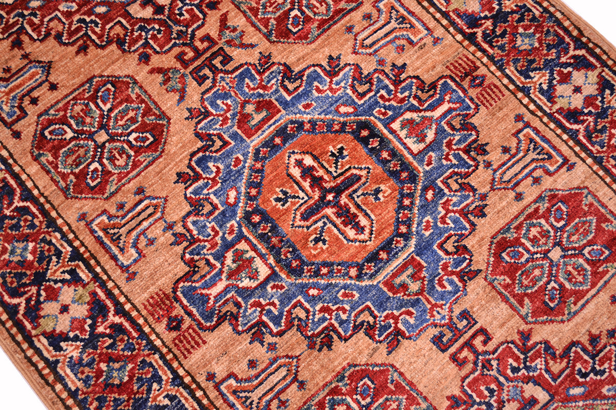 A close-up of a traditional patterned Kazak runner with a central medallion, featuring red, blue, and orange motifs on a warm peach background.