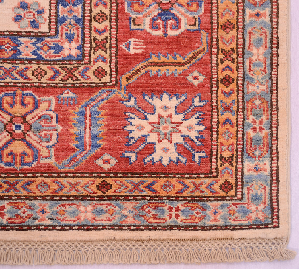 A close-up of a traditional handwoven fine Afghan Kazak carpet corner showing a red main border with cream, blue, and brown geometric and floral motifs, finished with a cream fringe.