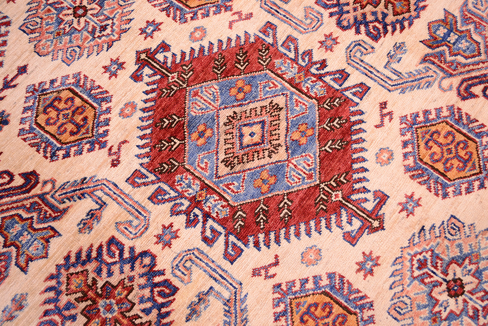 A close-up of a traditional patterned fine handmade Afghan Kazak carpet featuring a central red and blue medallion surrounded by geometric motifs on a light beige background.