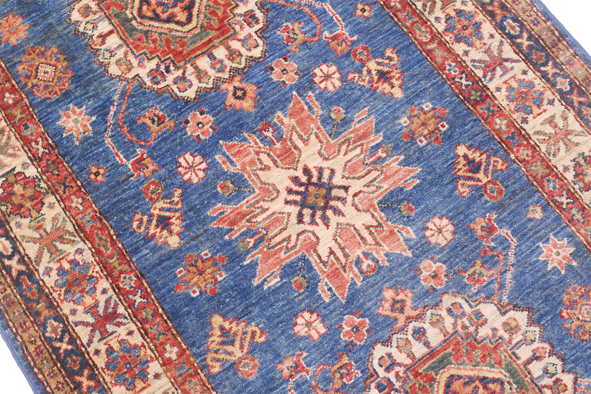 A close-up of a traditional patterned super kazak rug with a medium blue field, cream central medallion, and red, orange, and green floral motifs and borders.