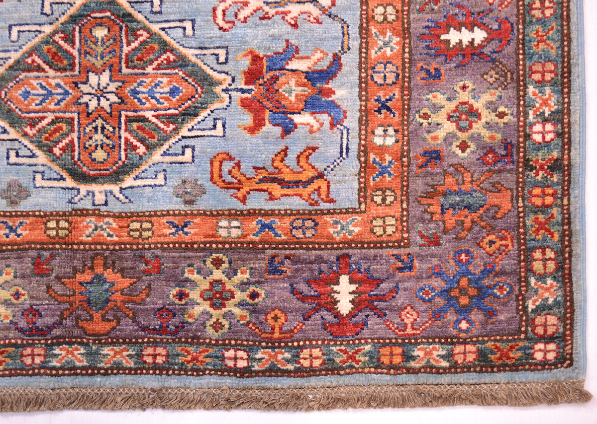 A close-up of an ornate patterned rug corner showing a blue field, rust and orange motifs, a mauve-purple border, and fringed edge.