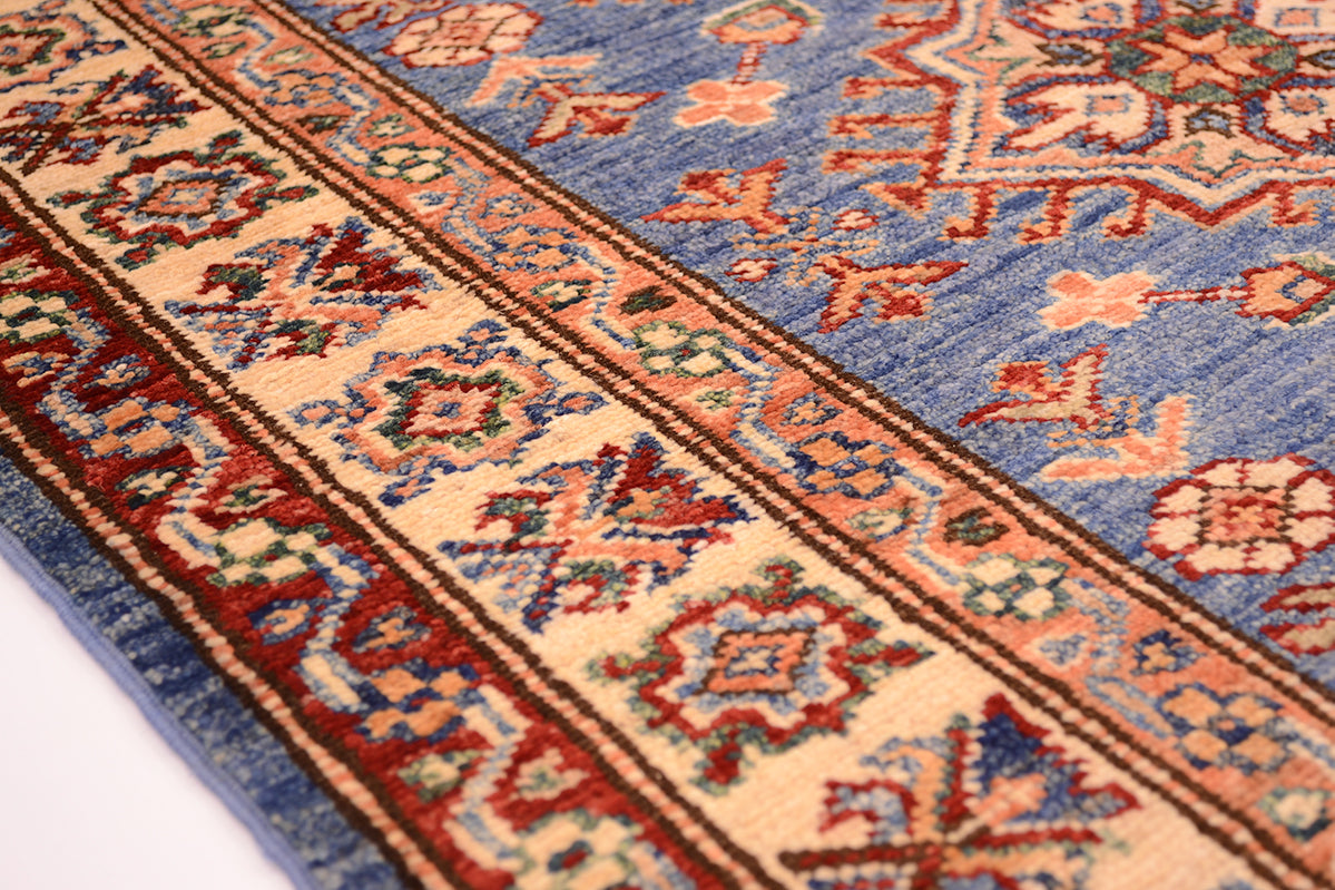 A close-up of a patterned handmade Afghan Kazak small rug showing a blue field with ornate floral motifs and a wide cream border with red, green, and brown decorative narrow borders.