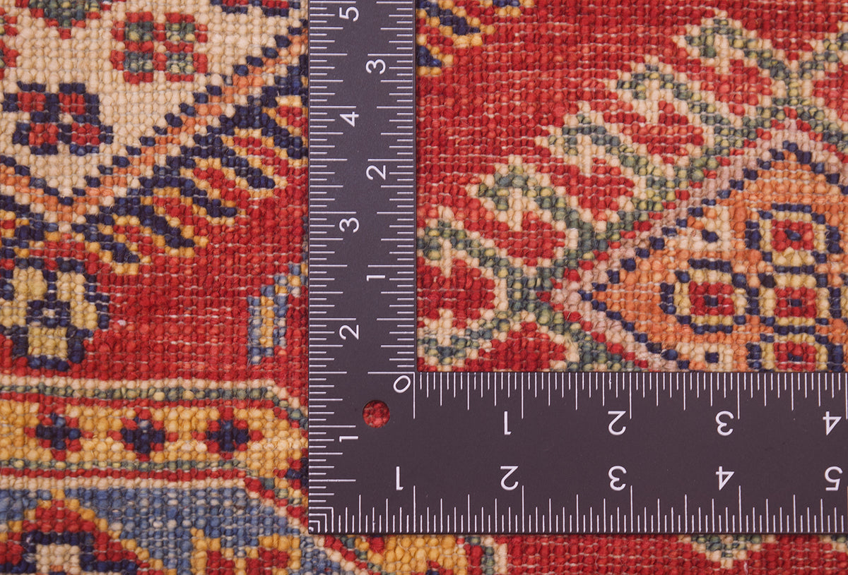Close-up of the hand-knotted back of a fine red Afghan Kazak Rug with a dark metal L-square ruler placed over it to show scale, showing a red, beige, and blue geometric design.