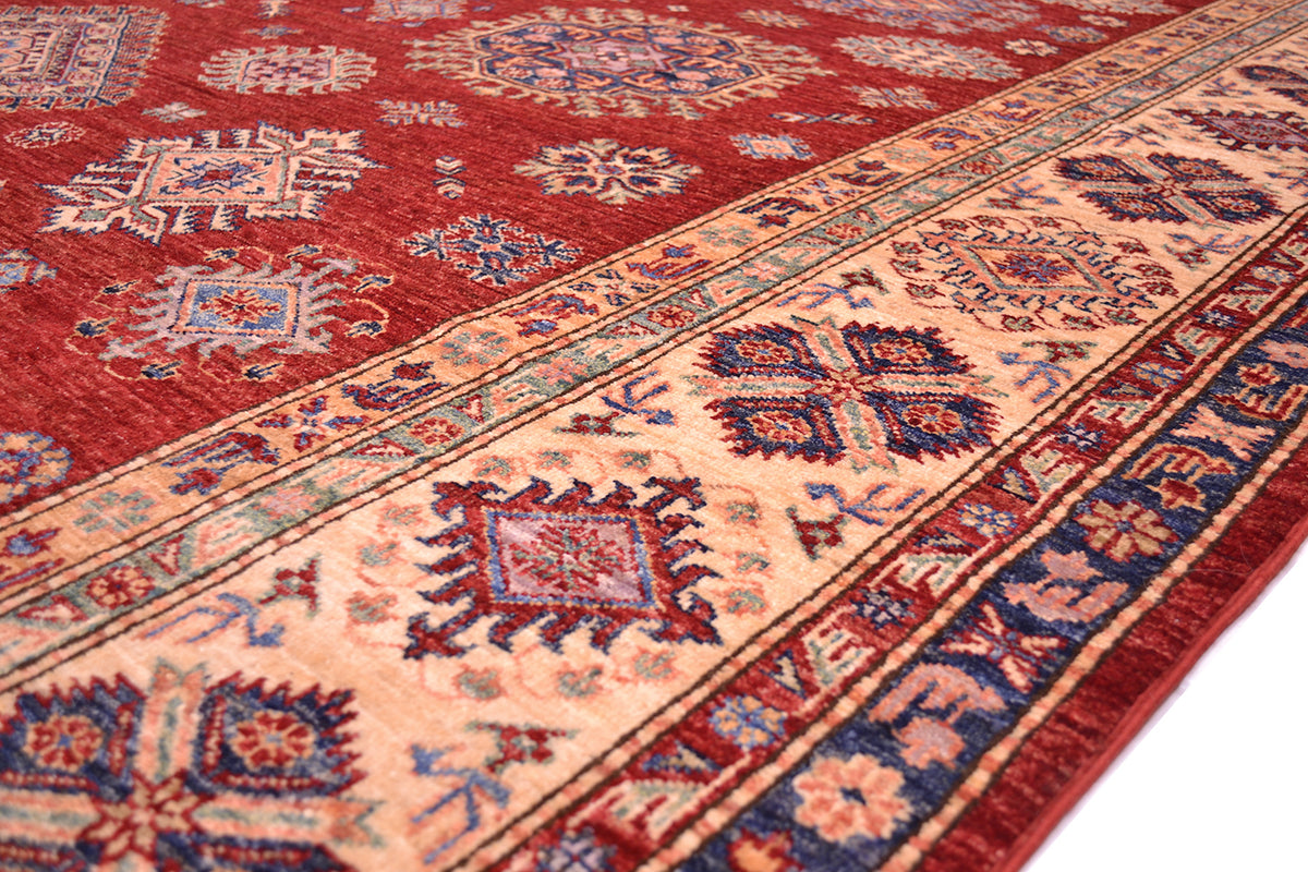 A close-up of a traditional fine red Kazak carpet with a deep red field, beige borders, and blue and orange accents.