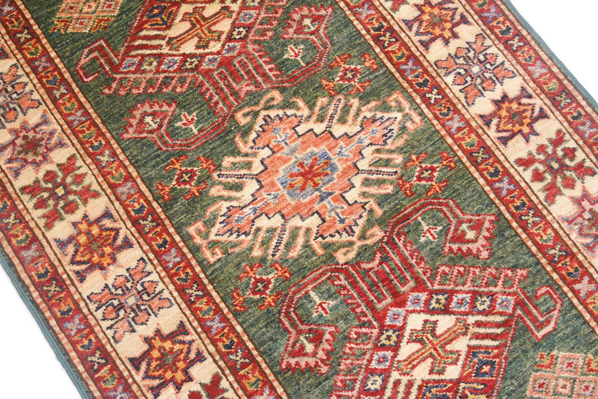A close-up of a traditional patterned Kazak Runner rug with a green field, red and beige medallions, and geometric borders.