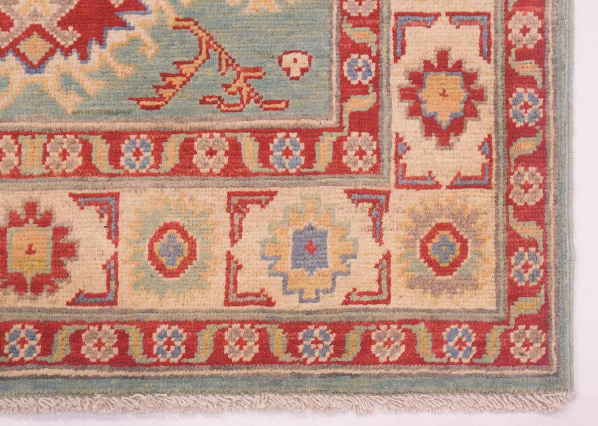 Close-up of a patterned Kazak rug corner showing a cream main border, red narrow borders, and a light blue-green outer edge with fringe.
