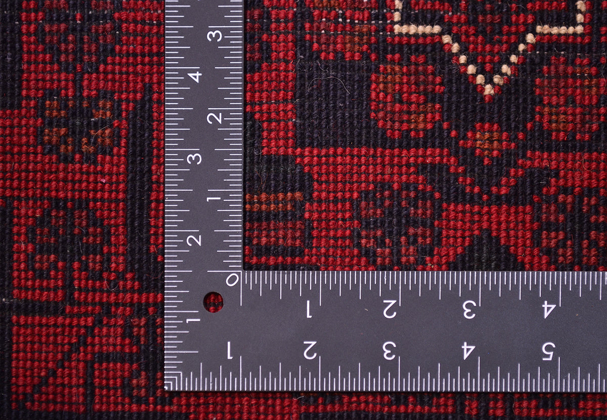 Close-up of a red and black patterned Afghan runner rug with a metal ruler placed over it to show scale, likely a rug or fabric swatch.