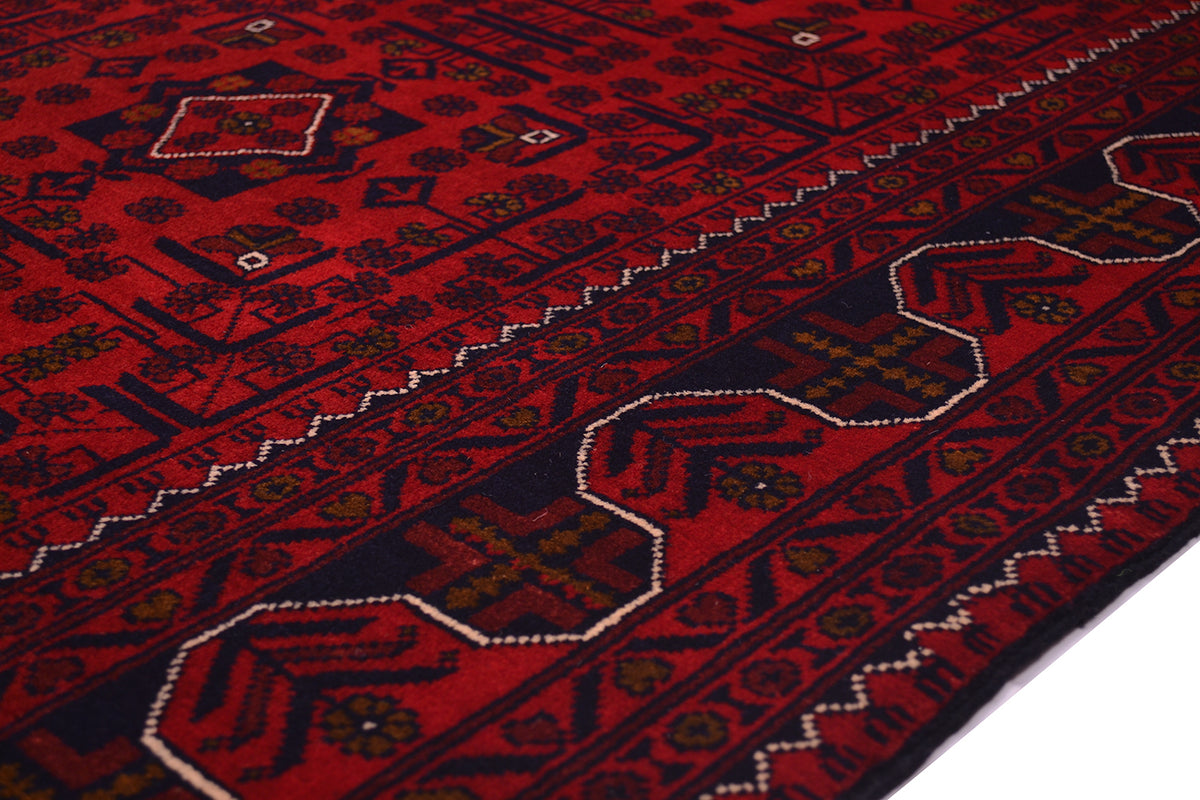 A close-up of a red patterned Afghan rug with dark navy and beige geometric borders and motifs.