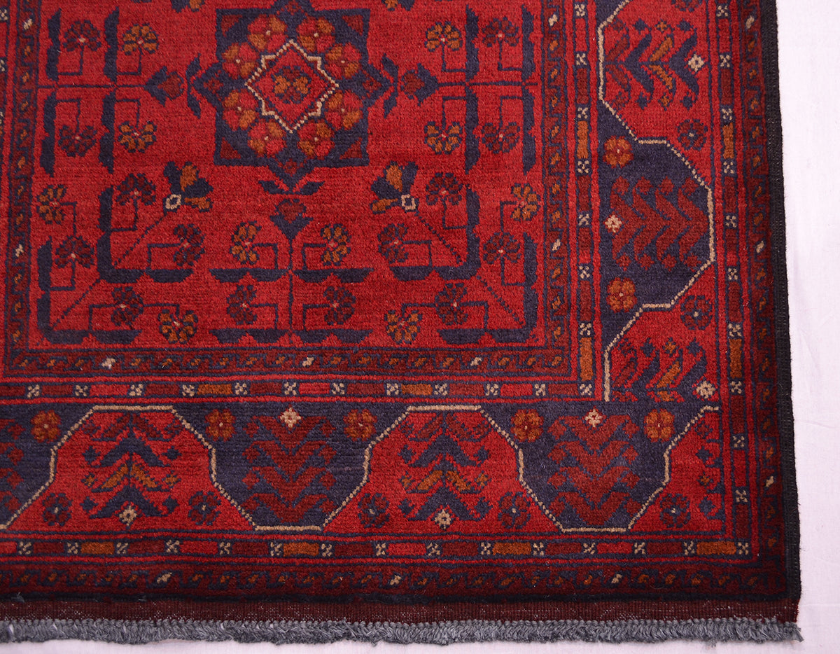 A close-up of a corner of a traditional afghan rug corner, featuring a deep red field with dark navy blue geometric borders and floral motifs in rust, beige, and orange.