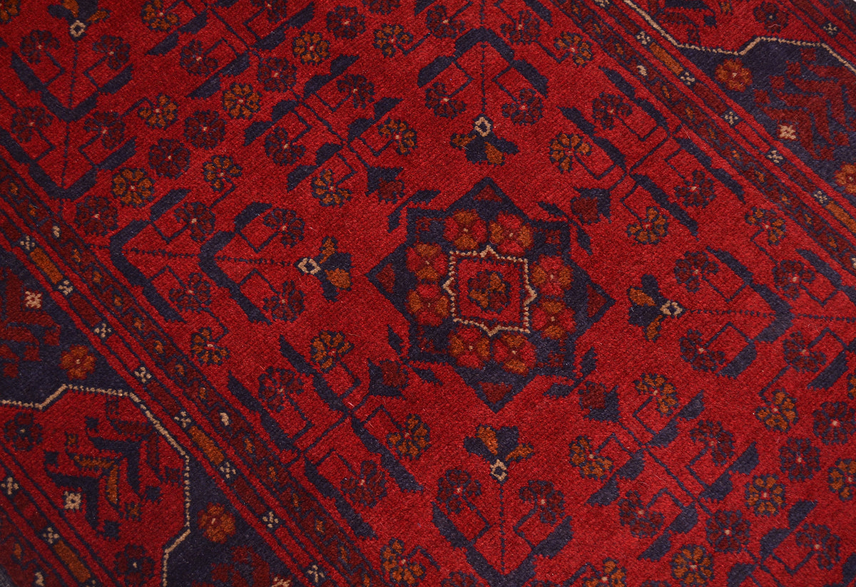 A close-up of an Afghan patterned runner rug with a deep red field, dark navy blue geometric borders, and small floral motifs in orange, beige, and white.