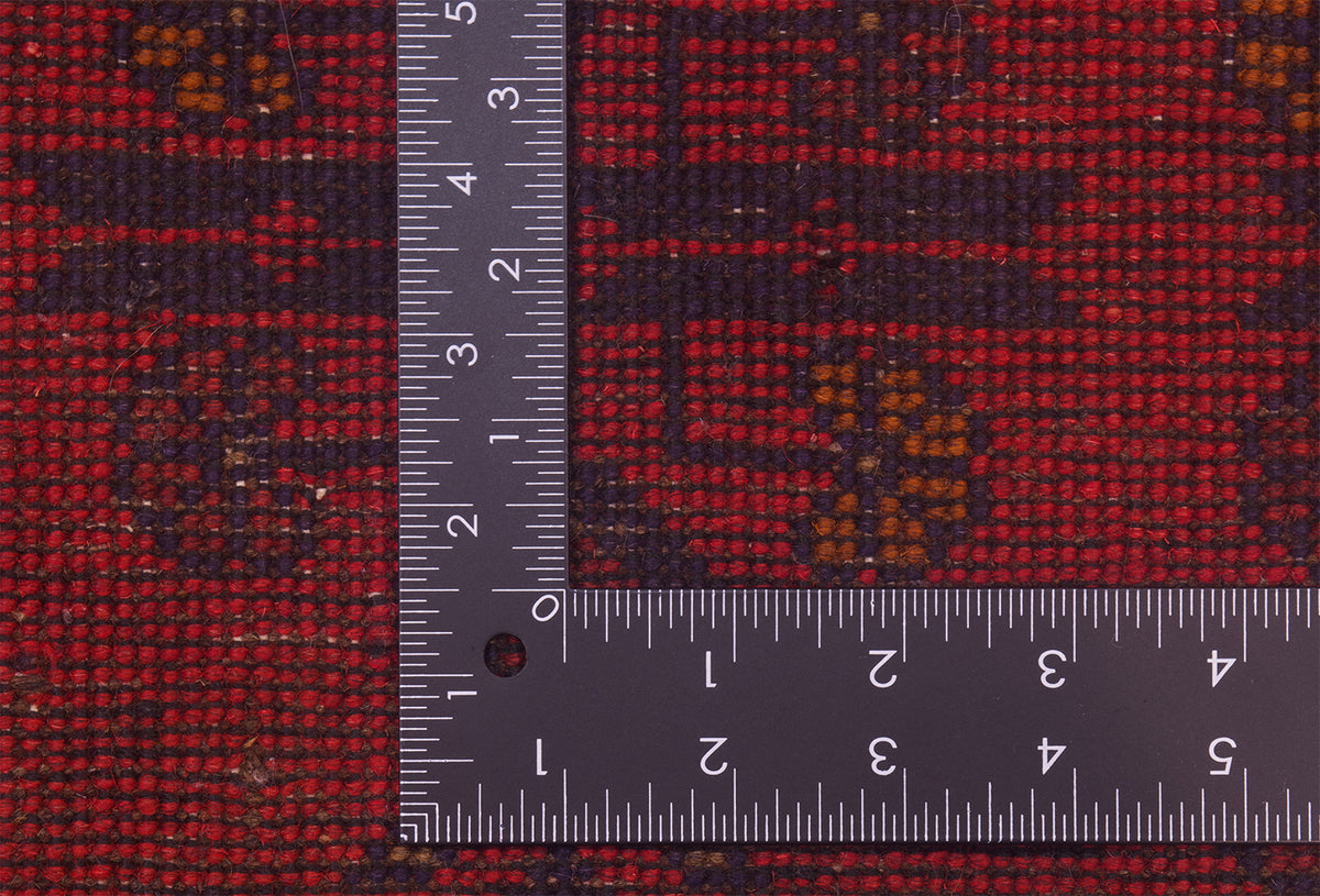 Close-up of back of a red patterned afghan rug with a dark ruler placed on top to show scale, highlighting the textile's weave and design.