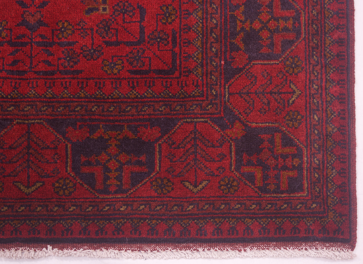 A close-up of a traditional afghan patterned rug corner, showing a deep red field with dark navy blue geometric medallions and borders, accented with small tan details and white fringe along the bottom edge.