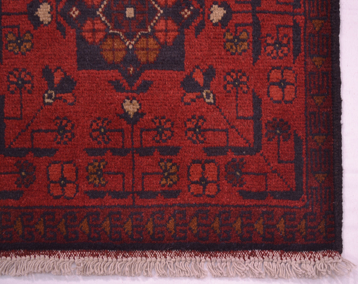 A close-up of an Afghan rug corner showing a deep red field with dark navy geometric and floral motifs, a patterned border, and light fringe.