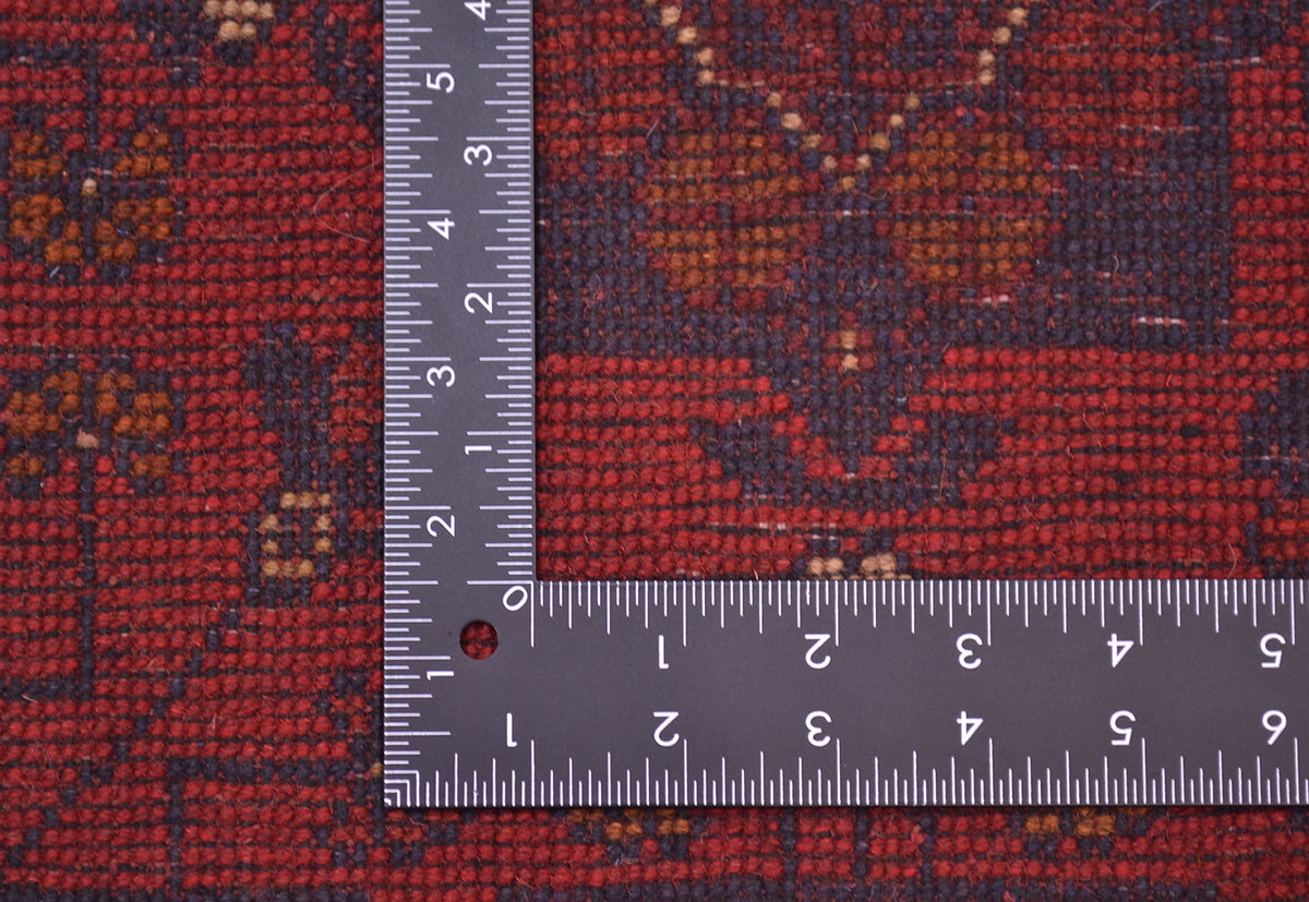 Close-up of back of a patterned with a metal L-square ruler placed on it to show scale. The runner has a dark red base with navy and brown geometric and floral motifs.