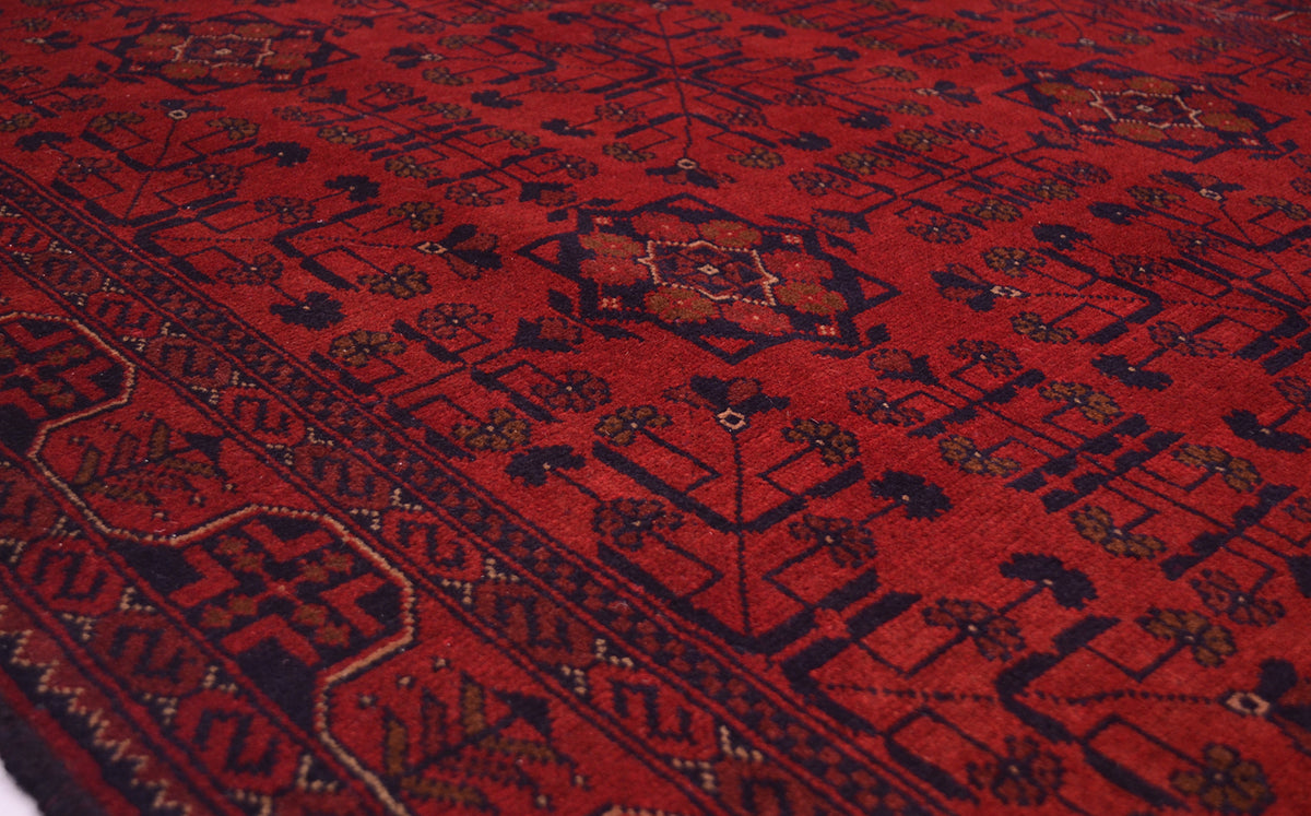 A close-up view of a traditional patterned handmade rug with a deep red field, dark navy geometric borders, and small floral motifs.