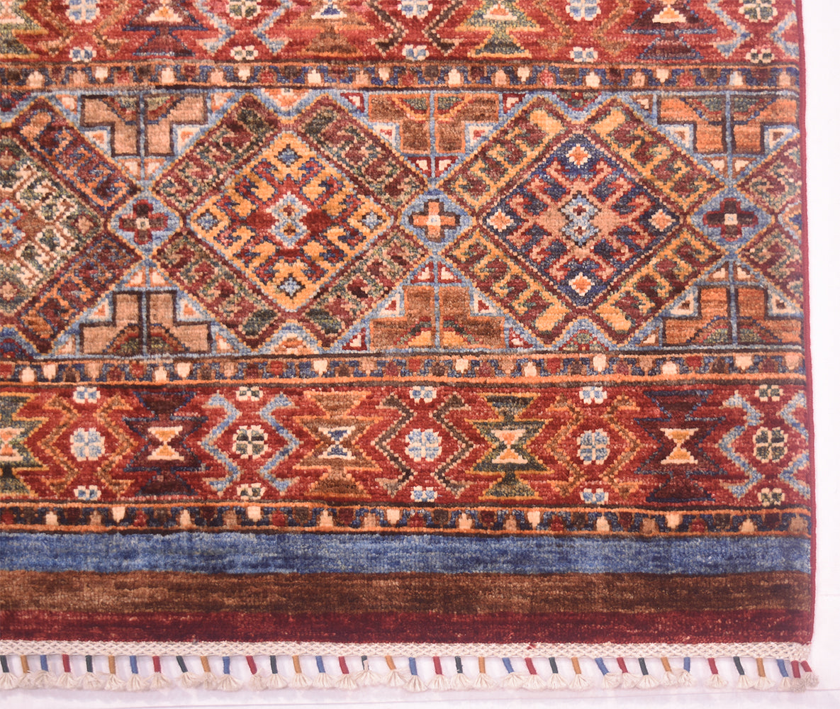 A close-up of a traditional patterned multicolour Afghan Khorjin rug with geometric diamond motifs in red, blue, beige, and brown, featuring a fringed edge.