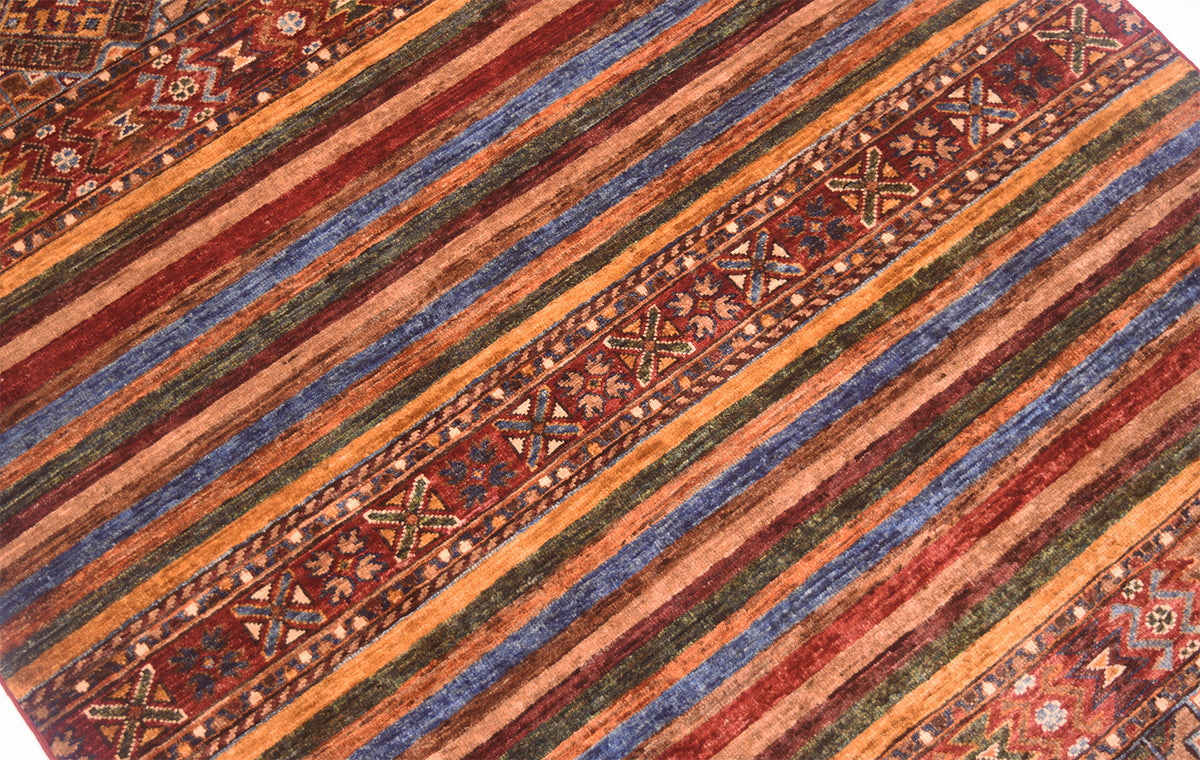 A close-up of a patterned Khurjeen rug with diagonal stripes in rust red, orange, blue, green, and brown, plus geometric border motifs.
