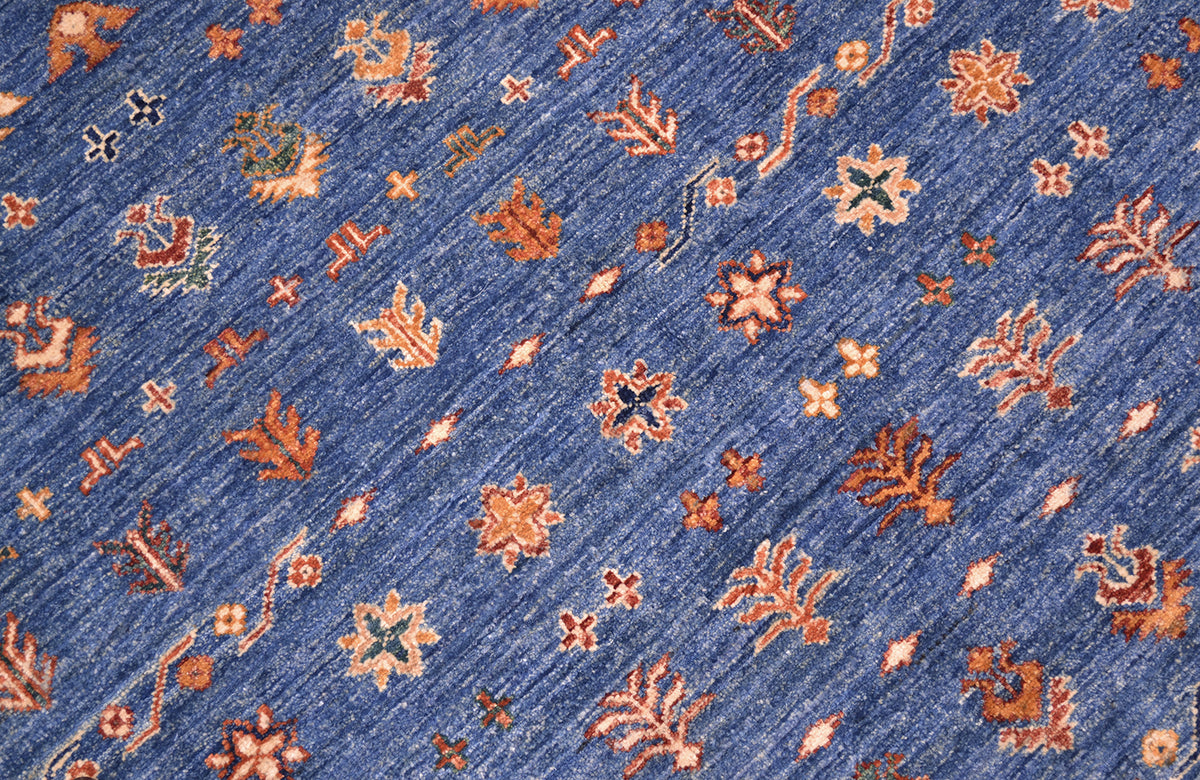 A close-up of a blue patterned Afghan Khorjin carpet with repeating floral and geometric motifs in rust orange, beige, and small green accents.