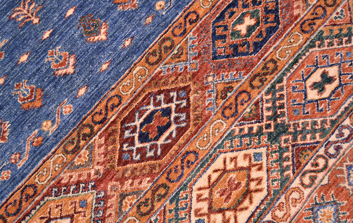 Close-up of a patterned Afghan Khorjin rug / carpet showing a blue field with small motifs and diagonal bands in rust orange, beige, navy, and green with larger geometric medallions.