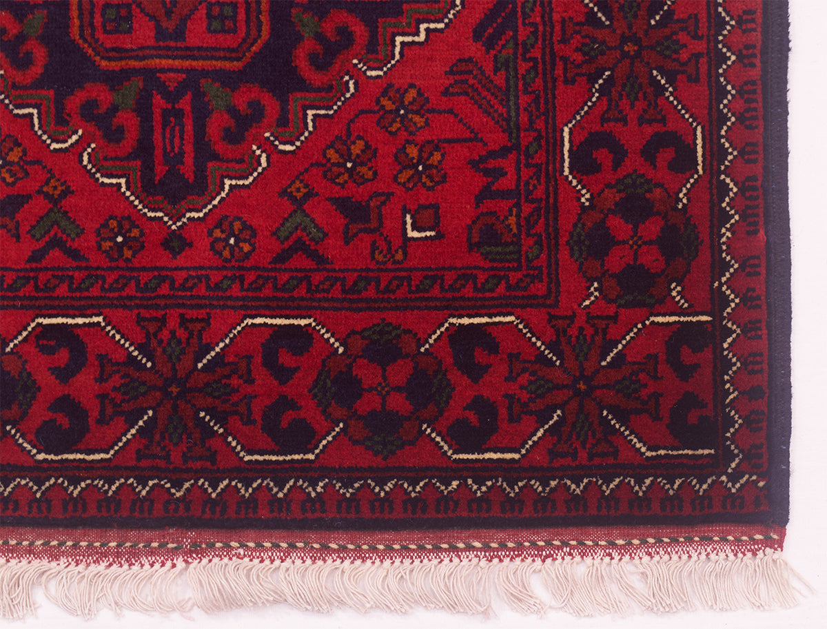 A close-up of a traditional patterned rug corner showing a deep red field with dark navy blue geometric medallions, floral motifs, and a decorative border, finished with cream-colored fringe.