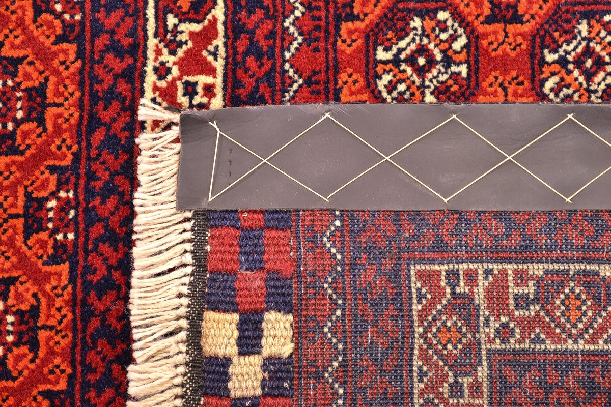 Close-up of back of a patterned Afghan runner rug corner showing cream fringe, a gray strapping attached to edge, and red, navy, and beige woven textile details.