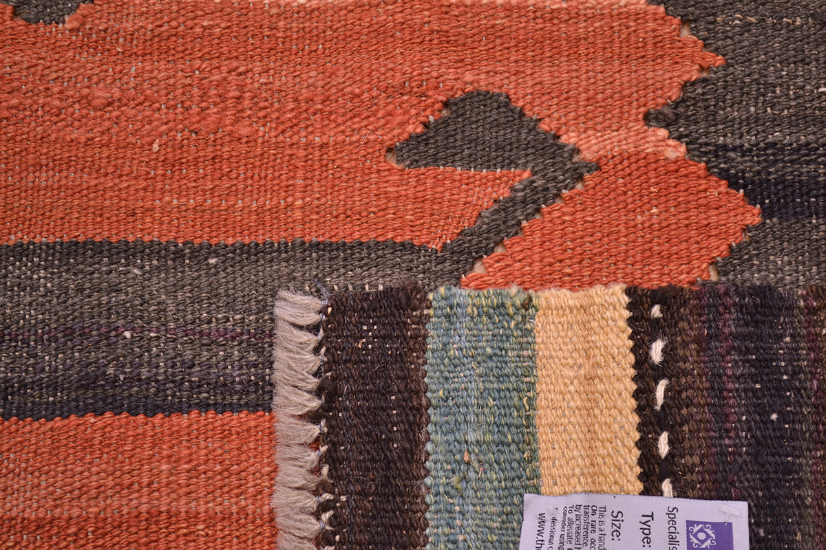 Close-up of the weave of a handwoven Afghan Labijar kilim rug showing a geometric pattern in terracotta orange, charcoal grey, and a striped section with beige, blue-green, and dark brown tones. A white product tag is visible in the lower right corner.