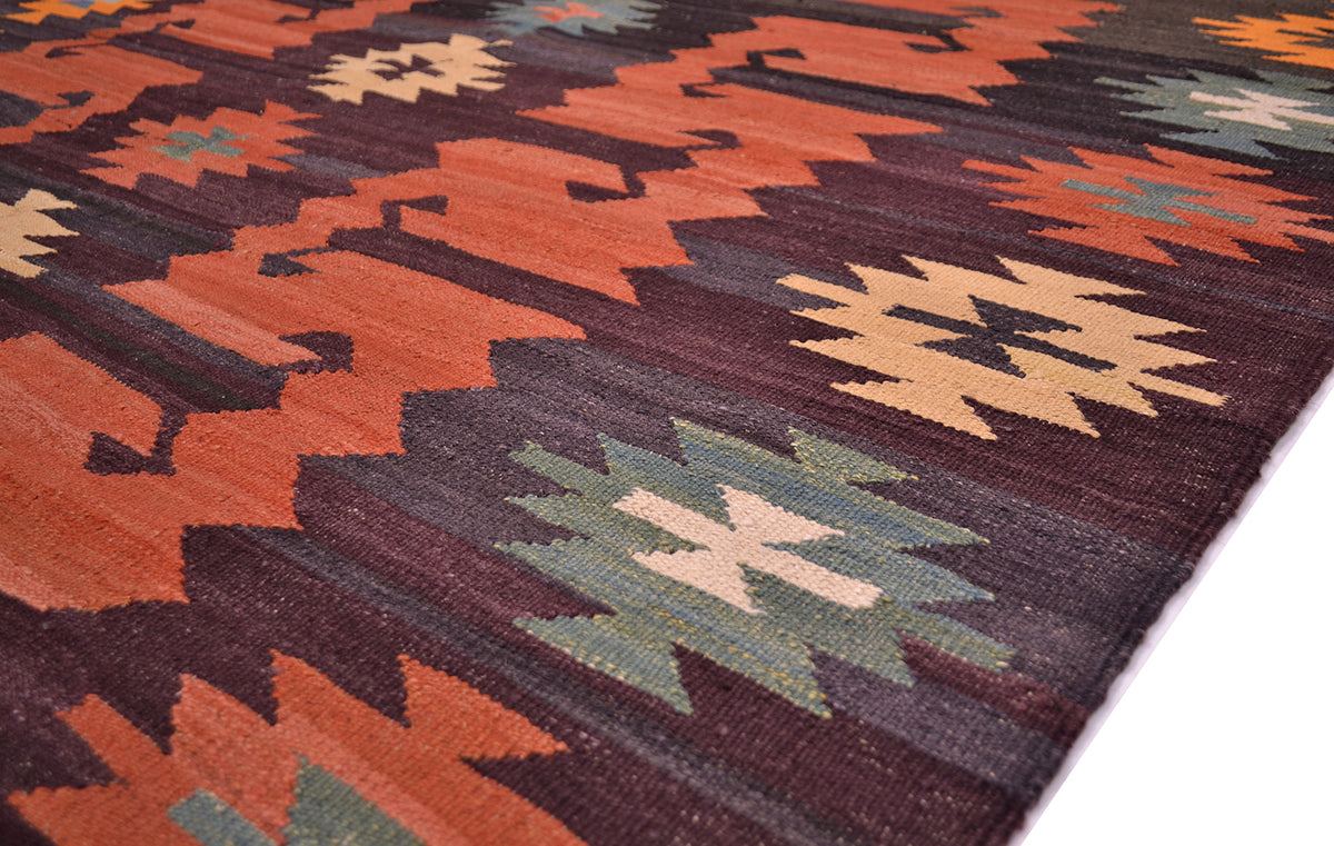 A close-up of a handwoven Afghan kilim rug with a geometric pattern in rust orange, dark brown, sage green, and beige, shown on a white surface.