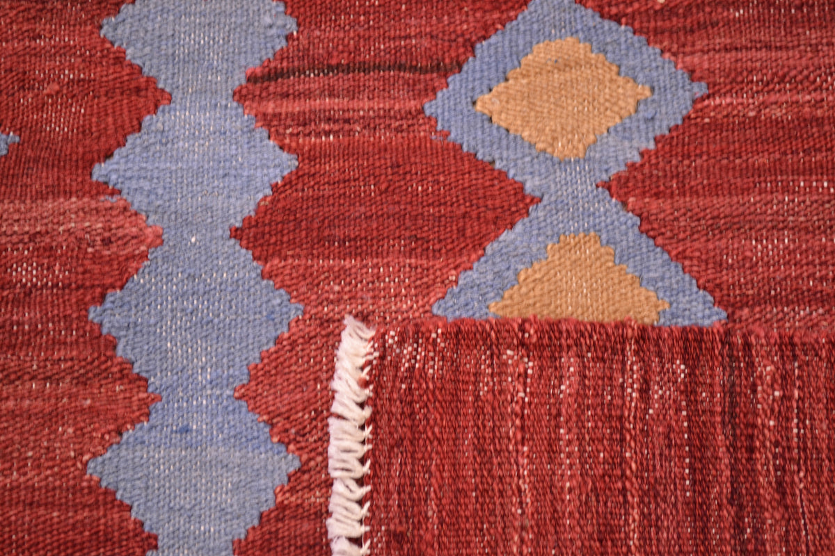 Close-up of the weave of a handwoven red Labijar Kilim 3x2m rug with a red field, light blue zigzag and diamond motifs, tan diamond accents, and a white fringed edge.