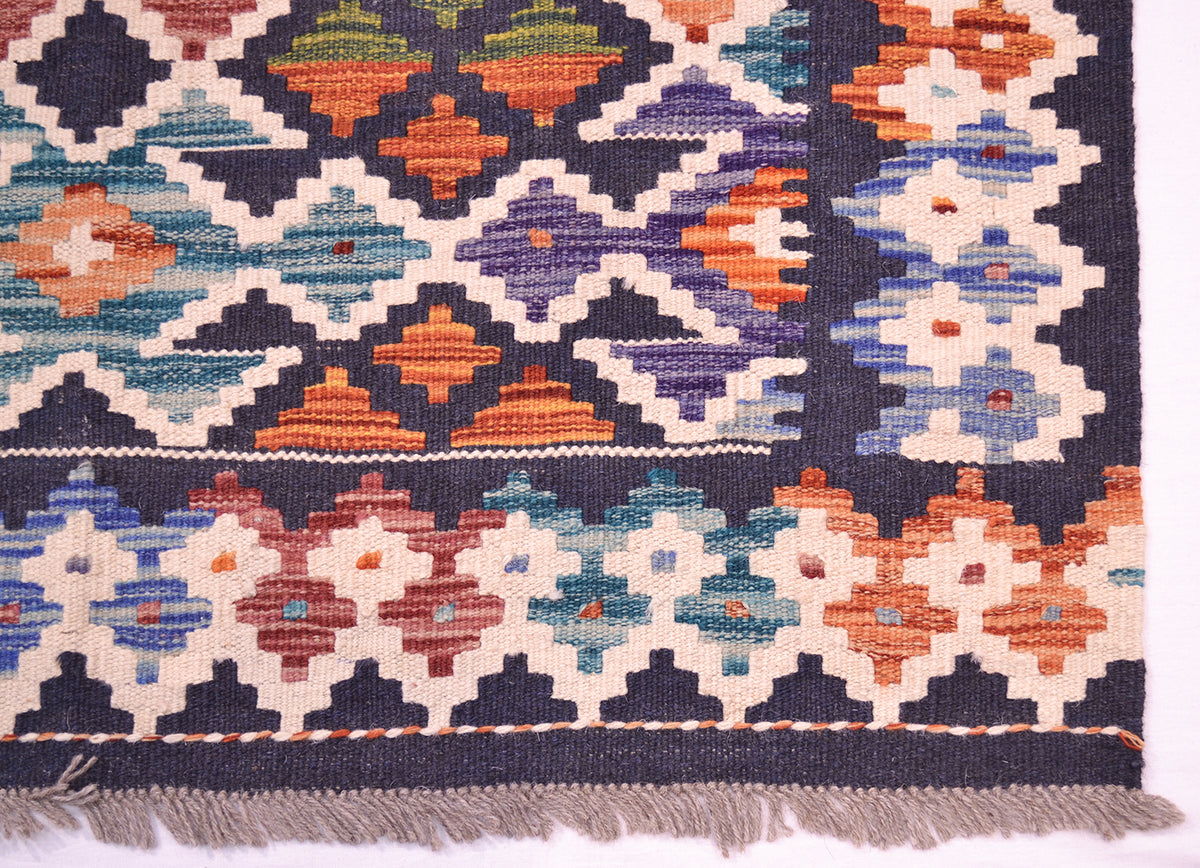 A close-up of a corner of colourful Kilim handwoven rug showing its geometric pattern and fringed edge. The design features a dark navy blue background with cream, orange, teal, purple, and red diamond and star motifs.